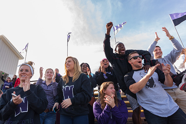 Group of students, some with painted faces, standing and cheering from the stands with purple & white and purple & black flags in the background